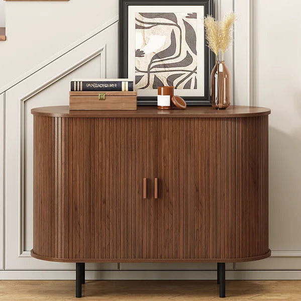 A curved wooden entryway cabinet with vertical fluting and black legs, styled in a modern home interior next to a staircase. The cabinet is decorated with books, a small vase, and a framed print.
