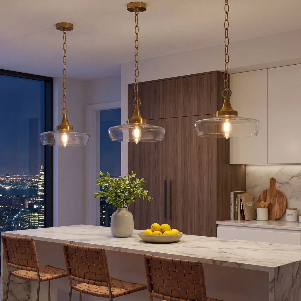 Three brass pendant lights with clear glass shades hang over a marble kitchen island in a modern kitchen setting.