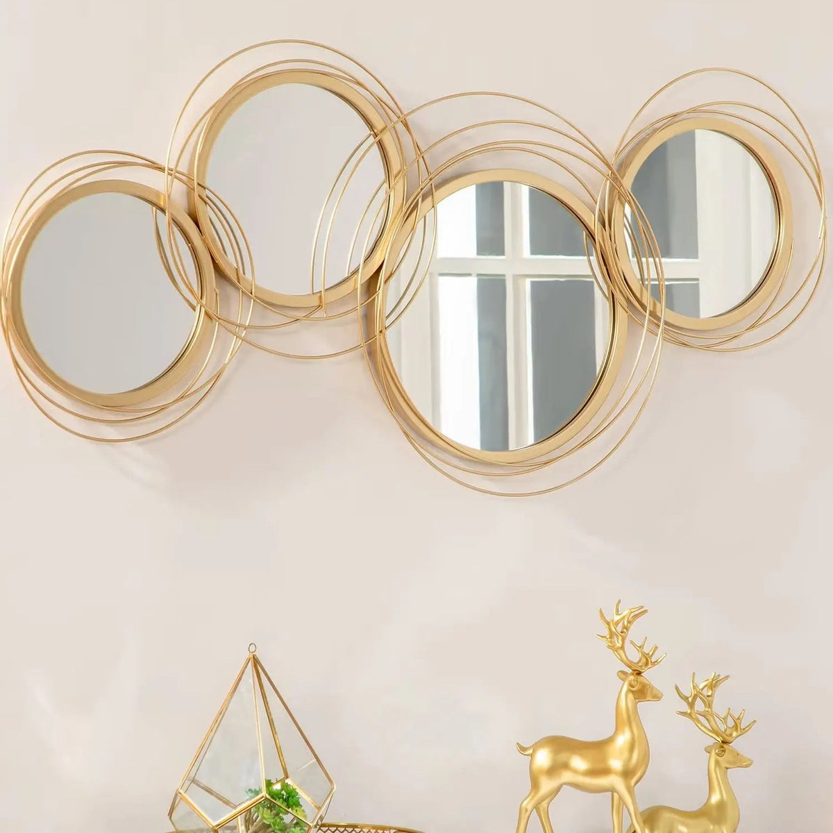 A set of five overlapping round wall mirrors with gold metal frames, mounted on a beige wall above a white console table decorated with gold deer figurines and a geometric glass terrarium.