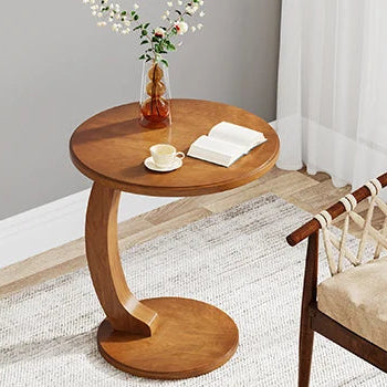 Wooden side table with a cup, saucer, and book next to a chair with woven backrest in a room.
