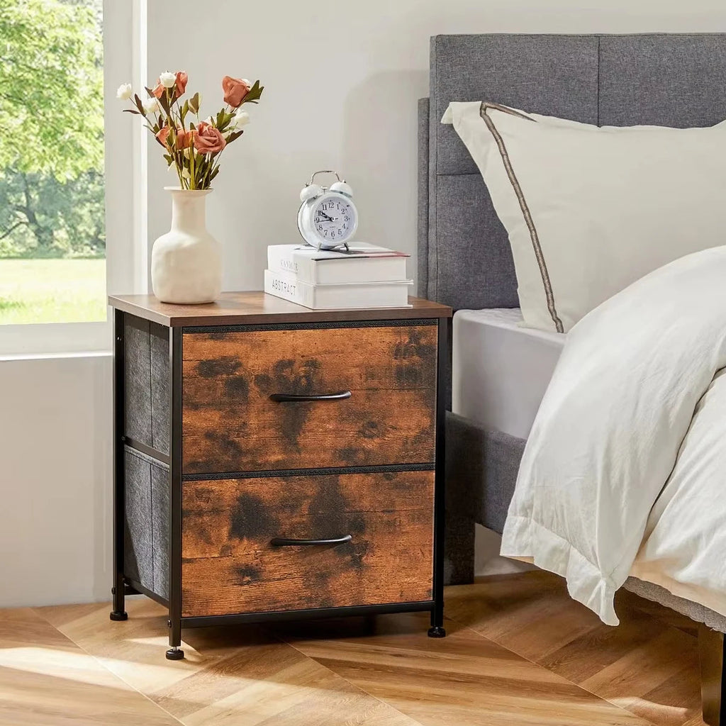 A rustic two-drawer nightstand with distressed brown wood fronts and dark gray fabric side panels, shown beside a bed in a bright bedroom. A vase with flowers, an alarm clock, and stacked books sit on top.