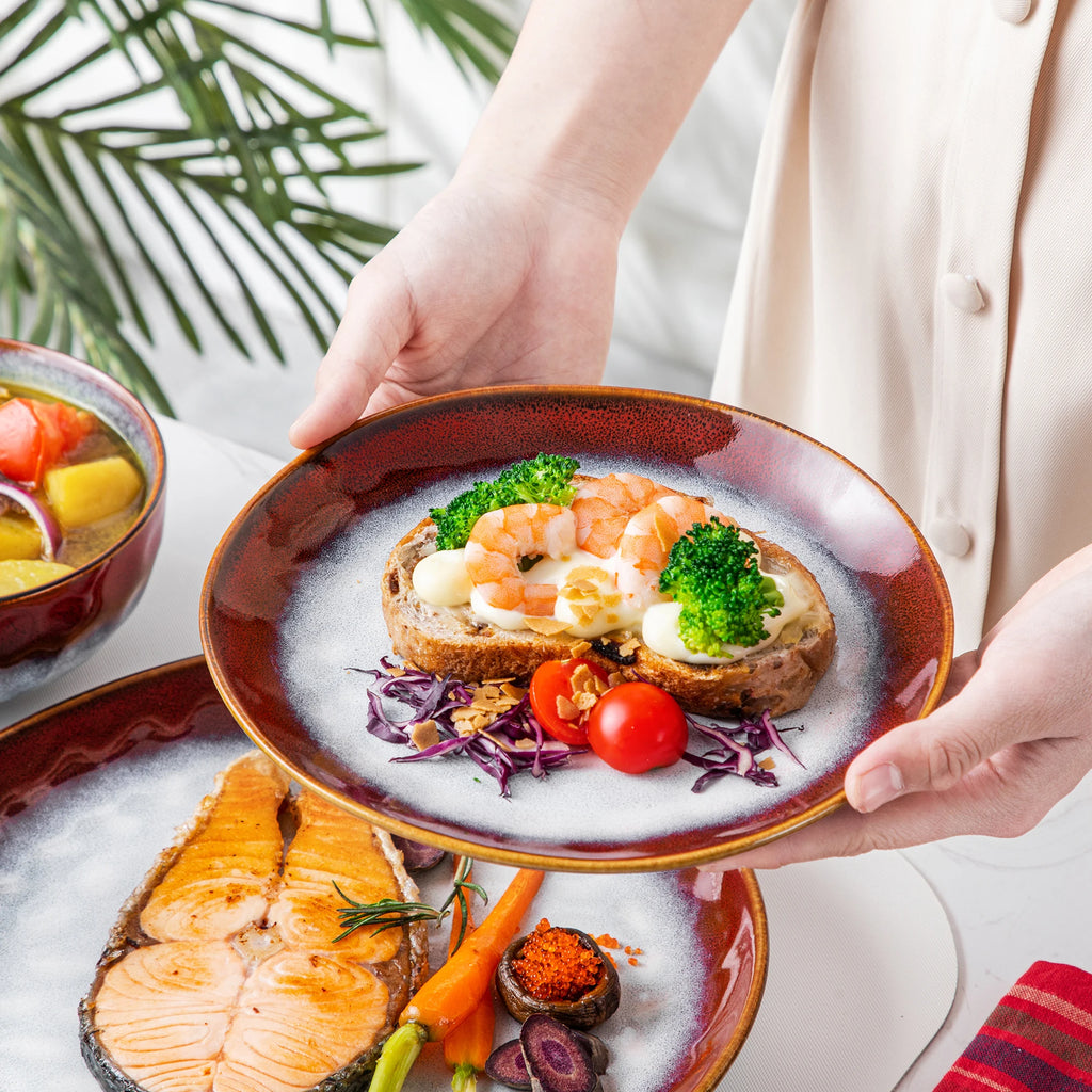 Hands holding a red-rimmed gray plate with shrimp toast and vegetables above a matching salmon plate.