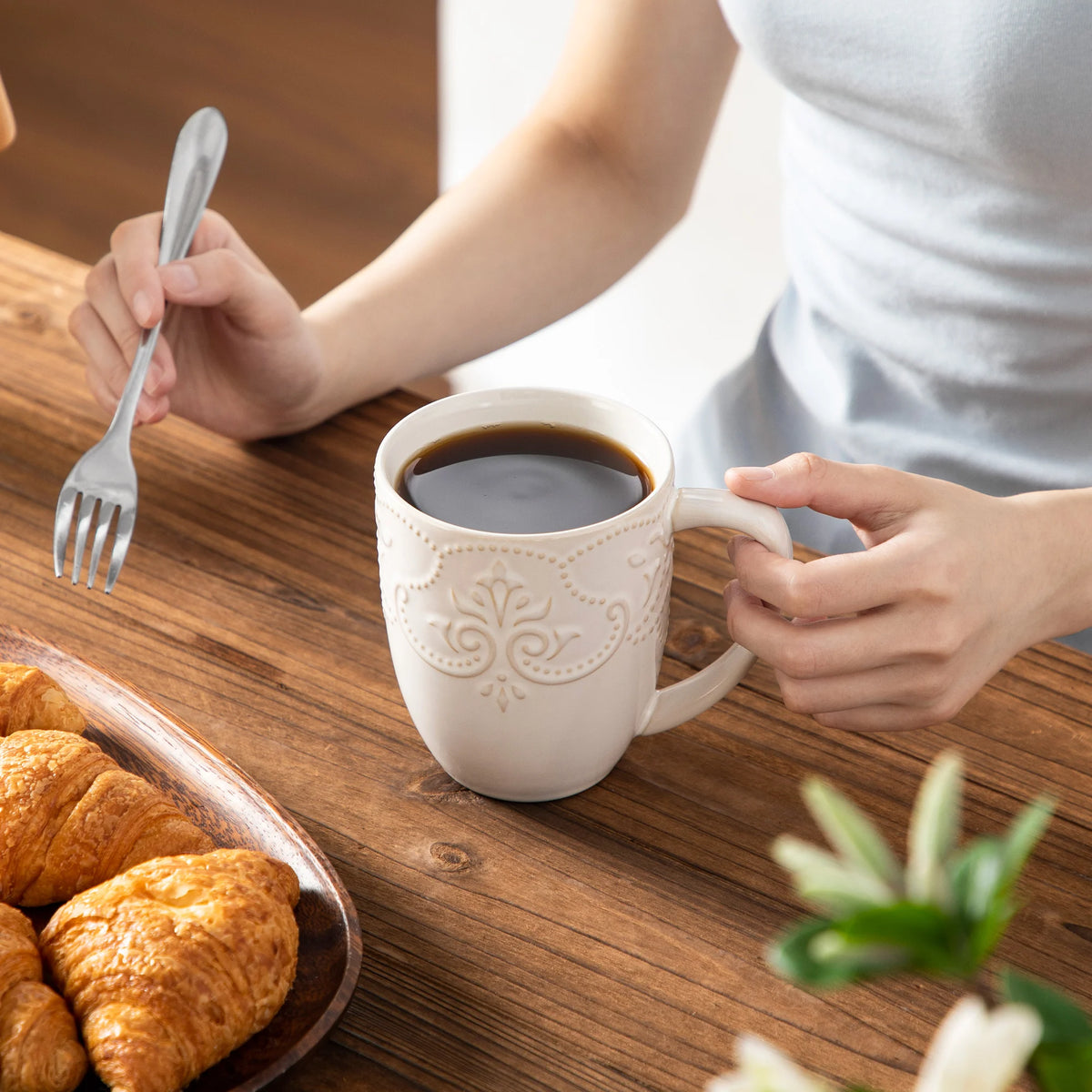 White GISELLE embossed mug filled with coffee, held at a wooden breakfast table beside pastries